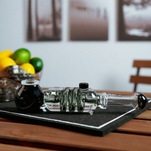 The Freeze Pipe, featuring a black mouthpiece and bowl, sits on a dark mat on a wooden table with lemons, limes, and blurred black-and-white photos visible in the background.