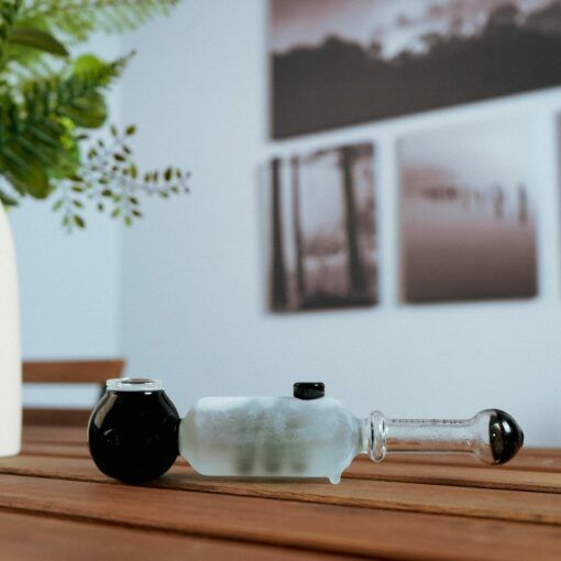 The Freeze Pipe, featuring black and frosted glass accents, sits on a wooden table. In the background are a white vase with greenery and black-and-white photos hanging on the wall.