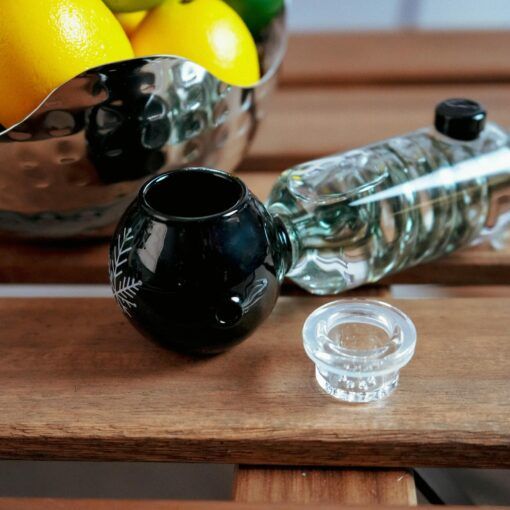 The Freeze Pipe, featuring a clear glass bottle with a black round cup and glass stopper, sits on a wooden surface. Behind it, a silver bowl contains yellow lemons and green limes.