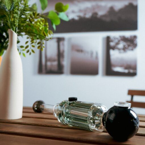 A Freeze Pipe Steamroller rests on a wooden table beside a cream-colored vase with green leaves, while out-of-focus black-and-white landscape photos are displayed on the wall in the background.