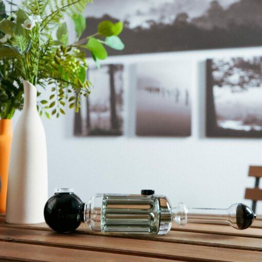 A Freeze Pipe Steamroller rests on a wooden table next to a white vase with green leaves and an orange bottle, with blurred black-and-white landscape photos hanging on the wall behind.