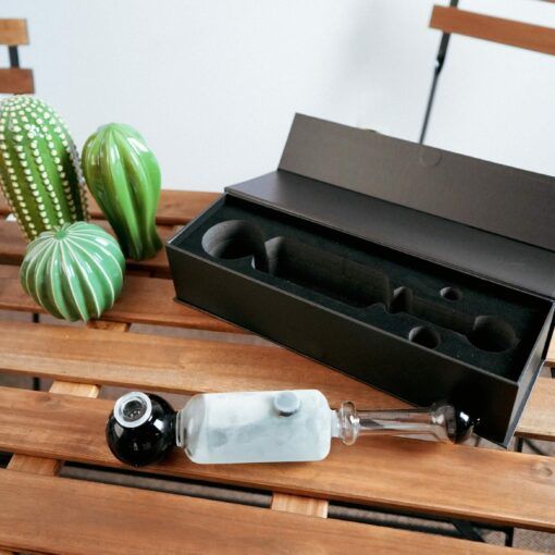 The Freeze Pipe Steamroller, featuring a black and clear design, sits on a wooden table beside a black box and two green ceramic cacti.