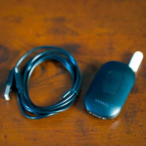 A black USB charging cable sits coiled beside the sleek Freeze Pipe Vape Pen, featuring a white mouthpiece and VESSEL branding, all resting on a wooden surface.