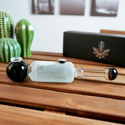 A Freeze Pipe Steamroller with black and frosted glass sections sits on a wooden table. Green decorative cacti, a black box with a white leaf, and blurred wall art create an inviting backdrop for this unique piece.