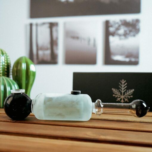 A Freeze Pipe Steamroller featuring black ends and a frosted white center sits on a wooden surface, with blurred wall art and green cacti visible in the background.