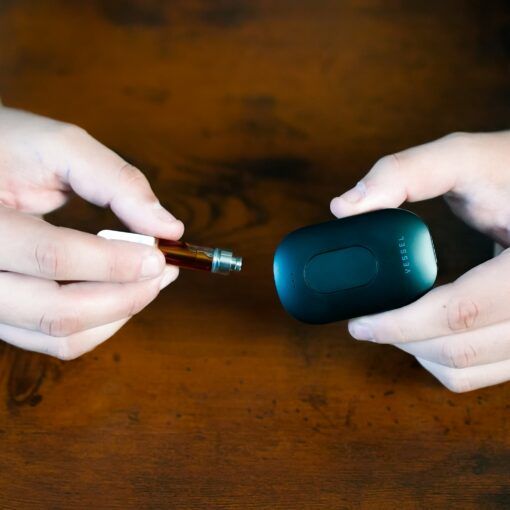 Two hands hold a Freeze Pipe Vape Pen and a vape cartridge filled with liquid against a wooden surface background.