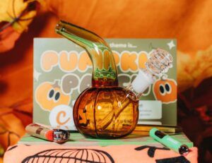 A pumpkin-shaped glass bong sits on a table with orange and green lighters nearby. In the background, a sign with cartoon pumpkins and partially visible text reads Pumpkin Patch, adding to the Cannabox autumnal vibe for October 2025.