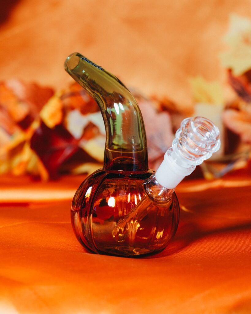 A small glass bong shaped like a pumpkin from the Cannabox October 2025 Pumpkin Patch sits on an orange surface, with autumn leaves blurred in the background.