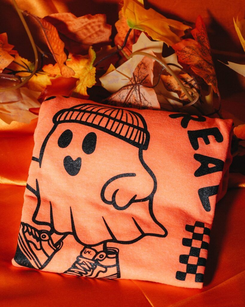 A folded orange shirt from the Cannabox October 2025 unboxing features a cute ghost in sneakers and a beanie, with "REAL" printed beside it. The shirt rests on orange fabric surrounded by artificial autumn leaves, perfect for a Pumpkin Patch vibe.