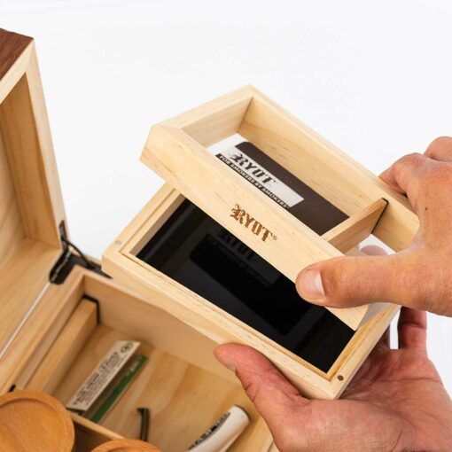 A person lifts a RYOT-engraved compartment from the HUMIDOR WALNUT COMBO BOX (8X11), designed for cigar storage, holding rolling papers, accessories, and a brown RYOT HERB GRINDER CARD pack.