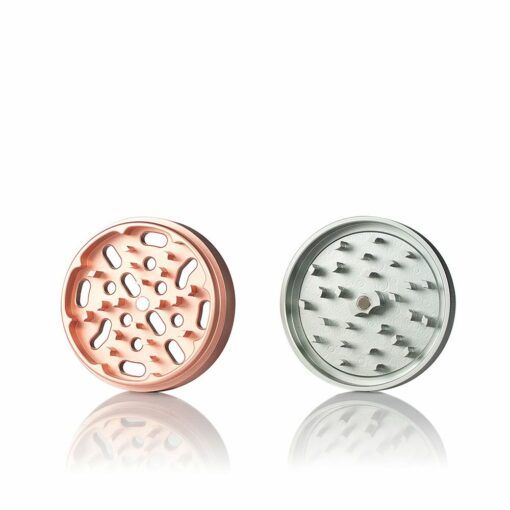 Two circular parts of the RYOT 4PC SOLID BODY GRINDERS—a rose gold piece with oval holes and a silver one with pointed teeth—are shown side by side on a white background, highlighting their premium metal build.