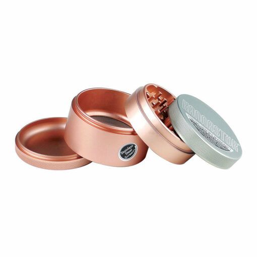 A rose gold RYOT 4PC SOLID BODY GRINDER with three compartments is shown open, displaying its teeth and storage sections. The lid, marked with "RYOT", is leaning against the grinder.