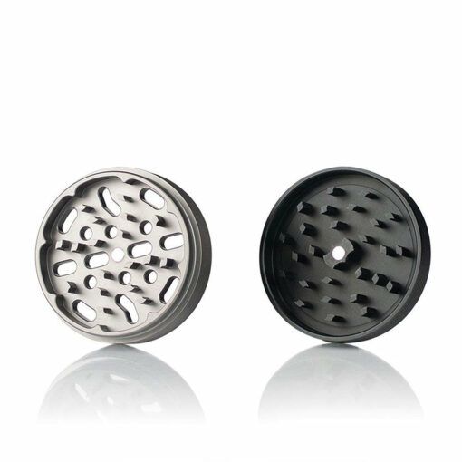 Two circular metal parts from the RYOT 4PC JAR BODY GRINDERS are shown side by side on a white background: the left is silver with oval holes, the right is black with sharp, angled teeth and a center hole.