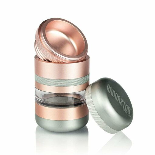 The RYOT GR8TR JAR BODY GRINDER features a metallic and glass body with rose gold and silver accents, a clear middle section, and two lids—one upright, the other on its side showing “HANNASTOR” branding.
