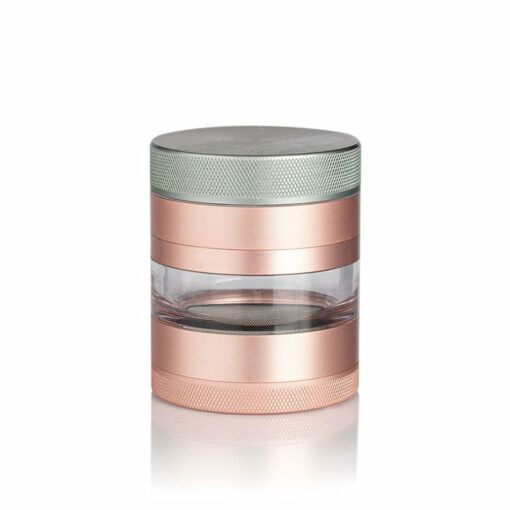 The RYOT 4PC JAR BODY GRINDER features a metallic cylindrical design with rose gold and silver accents, a transparent middle band for viewing the contents, and is displayed against a white background.