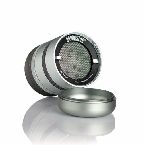 A RYOT GR8TR SOLID BODY GRINDER lies on its side with the lid off, exposing the mesh screen inside. The silver and black parts are reflected on a white surface.