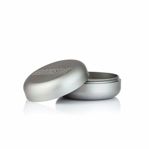 A round, silver RYOT GR8TR SOLID BODY GRINDER sits on a white background with its lid partially open to reveal the empty interior. Embossed text is visible on the top of the lid.