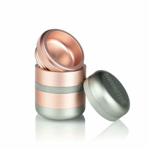 The RYOT GR8TR Solid Body Grinder is a metallic, cylindrical grinder with a rose gold and silver finish. Shown with its lid and top unscrewed to display the interior. The RYOT logo appears on the lid.