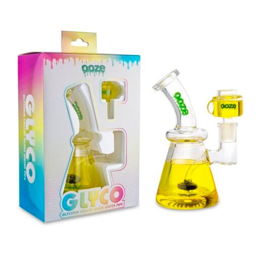 The Ooze Glyco Bong Glycerin Chilled Glass Water Pipe with yellow glycerin liquid is displayed next to its colorful packaging labeled GLYCO and ooze, featuring a window that reveals the unique glass pipe design.