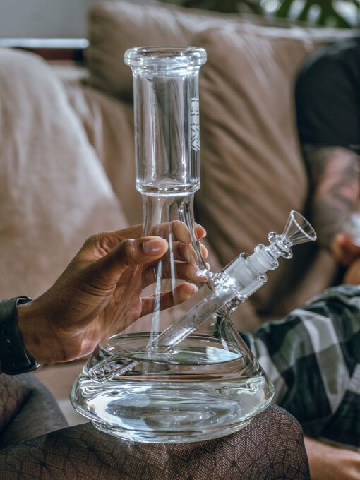 Someone holds a GRAV® Large Deco Beaker Bong filled with water while sitting on a couch, as another tattooed person relaxes in the background.