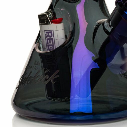 A close-up of the Gear Premium 12" Sidekick Beaker (Terminator Finish) Water Pipe with a built-in lighter holder, holding a white and red lighter. The beaker has dark, iridescent purple and blue reflections and "Sidekick" etched on the base.