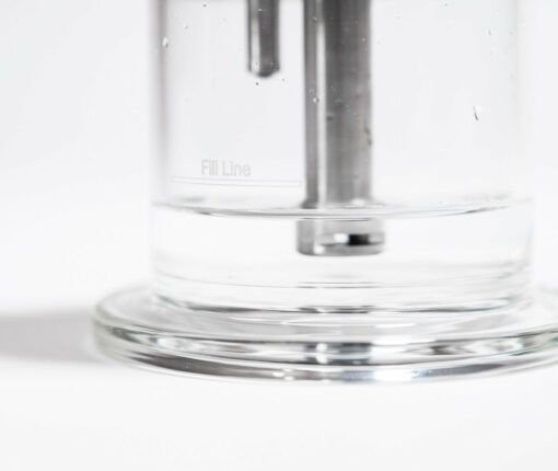 A close-up of the Trident V3, a transparent glass container with a visible fill line and some water inside. Metal rods are partially submerged, all set against a white background.