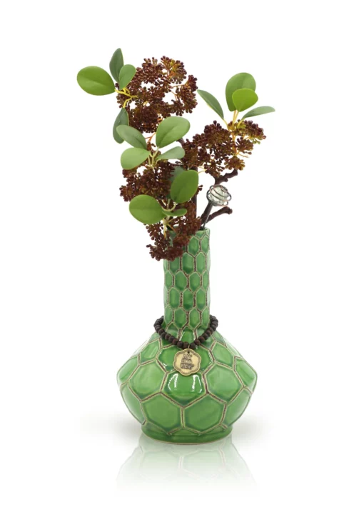 The TURTLE VASE BONG features a green honeycomb pattern, holding leafy stems and clusters of dark red flowers. A round gold medallion on a rope adorns its neck, set against a white background.