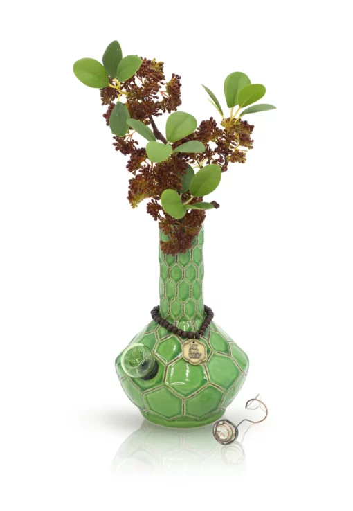 The TURTLE VASE BONG, featuring a green hexagonal pattern, holds leafy branches with clusters of small dark red flowers and is adorned with a black cord, gold charms, and a wire pendant.