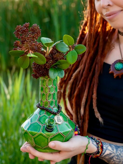 A woman with long dreadlocks holds a TURTLE VASE BONG featuring a green honeycomb pattern, filled with leafy branches and flowers. She wears colorful jewelry, standing against a lush green background.
