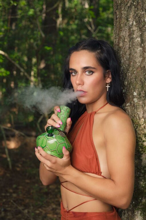 A person with long dark hair and a cross earring, in a rust-colored top, exhales smoke from the TURTLE VASE BONG while standing by a tree in a forest.