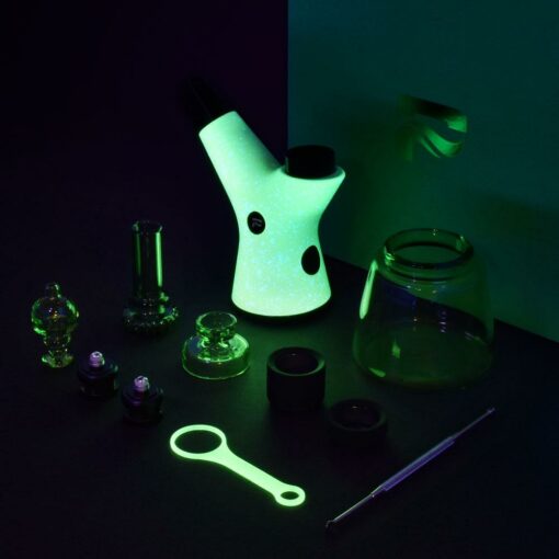 The Pulsar RöK Electric Dab Rig - Luna Glow features a glowing speckled design, multiple detachable parts and accessories—like glass chambers, caps, and a metal tool—displayed on a dark surface under green light.