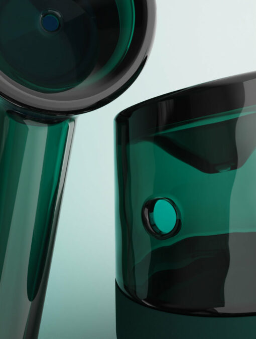 Close-up of Forge [Emerald]: two glossy, emerald-green glass objects with round, smooth shapes against a pale background. One upright and cylindrical; the other partially visible and angled.