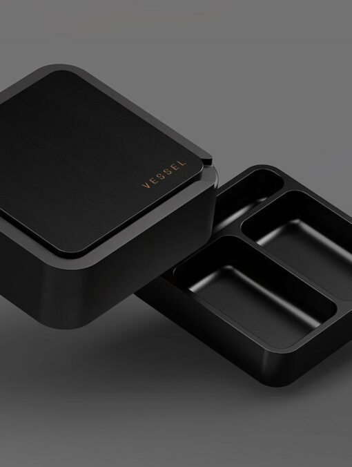 The Ash [Black] lunchbox, featuring the VESSEL logo on the lid, is shown partially open to display its three compartments against a simple gray background.