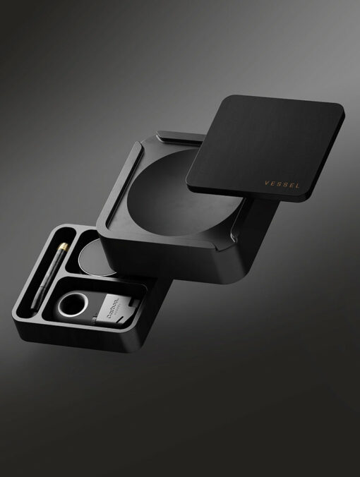 A sleek, black, square modular box labeled Ash [Black], opened to reveal compartments neatly holding a pen, lighter, and accessories—all arranged on a dark gradient background.