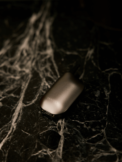 The Compass [Gunmetal] +, a metallic oval device with a sleek finish, sits atop a dark marble-patterned surface with white veins, evoking the look of a contemporary compass.