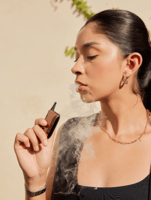 A woman with long dark hair pulled back exhales vapor while holding the Compass [Copper]+ vape. She wears gold jewelry and a sleeveless black top, sunlight casting compass-shaped shadows on the beige background.