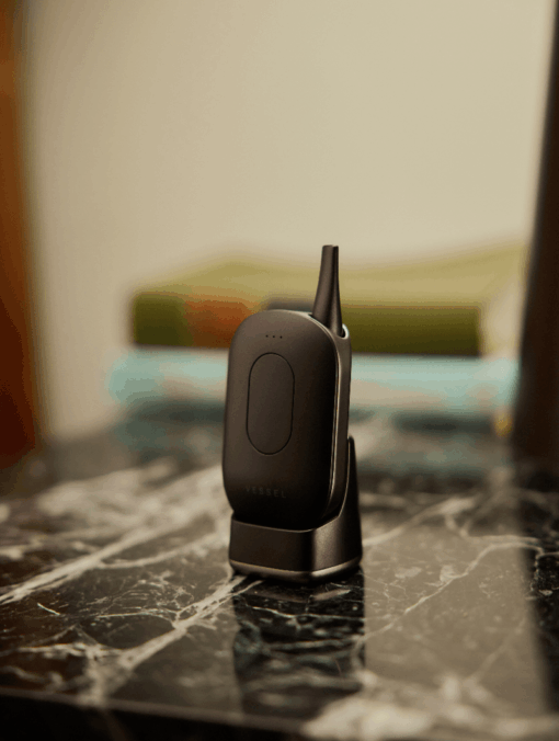 A Compass [Gunmetal] + device with an antenna stands upright in its charging dock on a marble surface, with blurred books visible in the background.