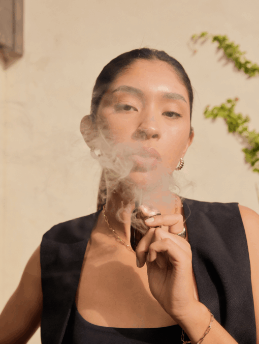 A woman wearing gold and Compass [Copper] + jewelry and a dark sleeveless top exhales vapor from an e-cigarette, with smoke partly obscuring her face. Sunlight filters in, revealing a wall adorned with climbing plants in the background.