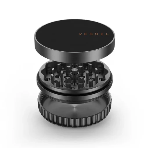 A gunmetal three-piece metal herb grinder with a partially removed lid reveals sharp grinding teeth. "VESSEL" is printed in orange on top. Complements the Drift Case [Gunmetal]. Set against a white background.