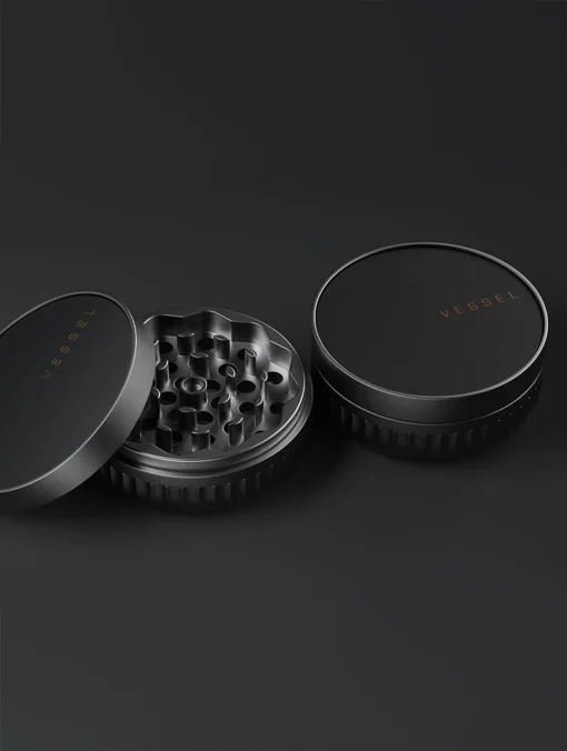 Two black circular grinders with textured edges—one open to show sharp teeth, both featuring VESSEL in gold on their lids—sit against a dark background next to the sleek Drift Case [Gunmetal].
