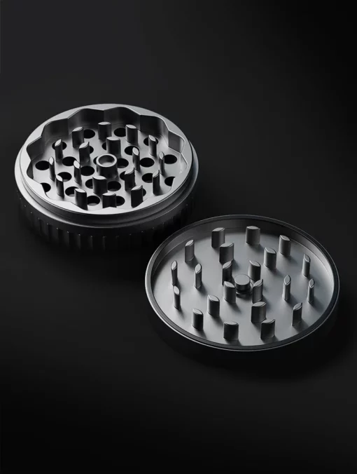 The Drift Case [Gunmetal] herb grinder is shown open on a dark background, revealing its sharp raised teeth and signature circular design on the interior grinding surfaces.