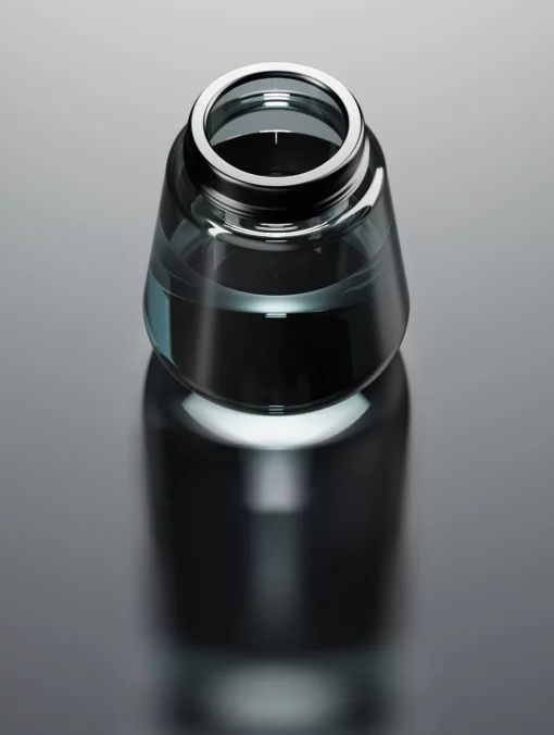 A close-up of Element [Black], a small clear glass ink bottle with a wide opening and black liquid, sits as a striking element on a reflective gray surface.