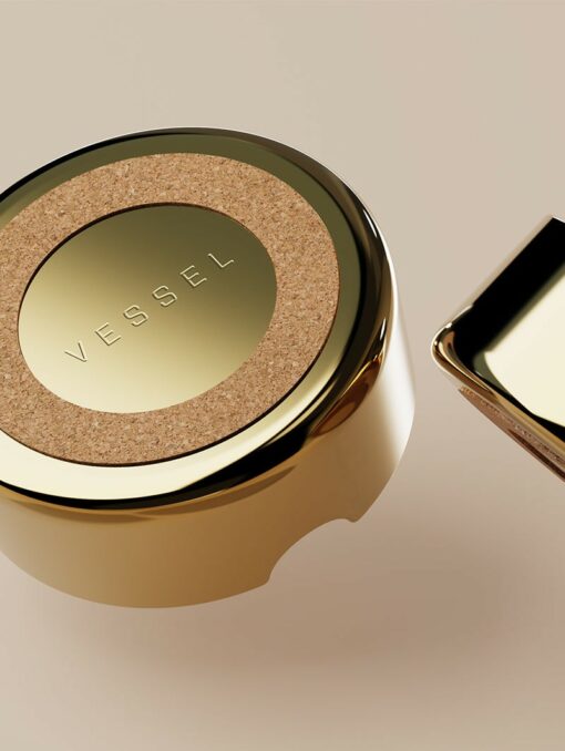 The Ember [Gold] is a round, gold-colored container lid with a cork center and VESSEL engraved on it, glowing like embers, displayed on a beige background beside a matching gold object.