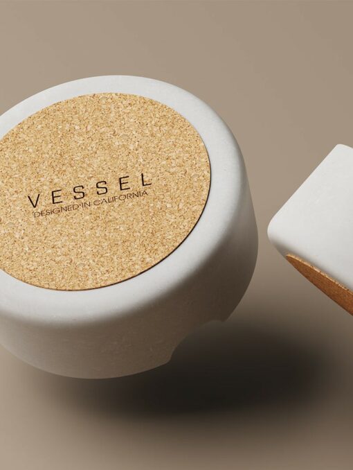Minimalist shot of Ember [Concrete]: a round white container with cork lid labeled "VESSEL DESIGNED IN CALIFORNIA" and part of a rectangular concrete piece featuring an ember-toned cork detail, set on a beige background.