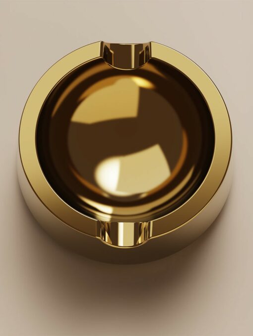 The Ember [Gold] ashtray, featuring a shiny round shape with two cigarette grooves, sits elegantly on a light beige surface.