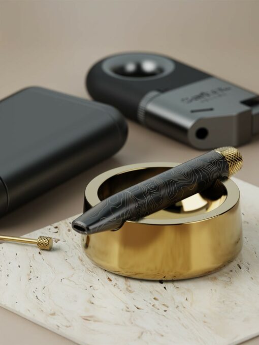 The Ember [Gold] vape pen, featuring a black and gold pattern, sits on a shiny gold ashtray with a lighter, dark case, and dab tool nearby—all arranged on a light marble surface.