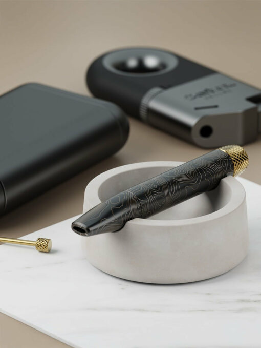 The Ember [Concrete] vape pen in black with gold accents sits in a white circular holder beside a vape cartridge tool. In the background, there’s a black lighter and matte black case on a marble and concrete surface.