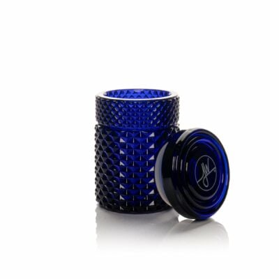 The Twenties Collection Jar | Cobalt, featuring a diamond-textured cobalt blue finish, sits open on a white background. Its matching lid displays a circular design with a white signature and rests beside the jar.