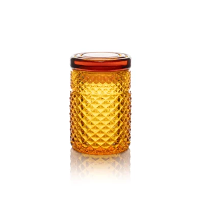 The Twenties Collection Jar | Amber is a small, amber-toned glass jar with a diamond pattern and matching lid, displayed on a white background with its reflection visible below.