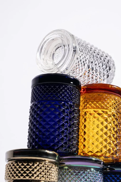 A stack of textured glass jars in clear, blue, gold, green, and amber against a white background, with the Twenties Collection Jar | Amber resting horizontally on top.